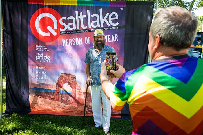 Man standing in front of a large magazine cover background while a man wearing rainbow takes his photo.