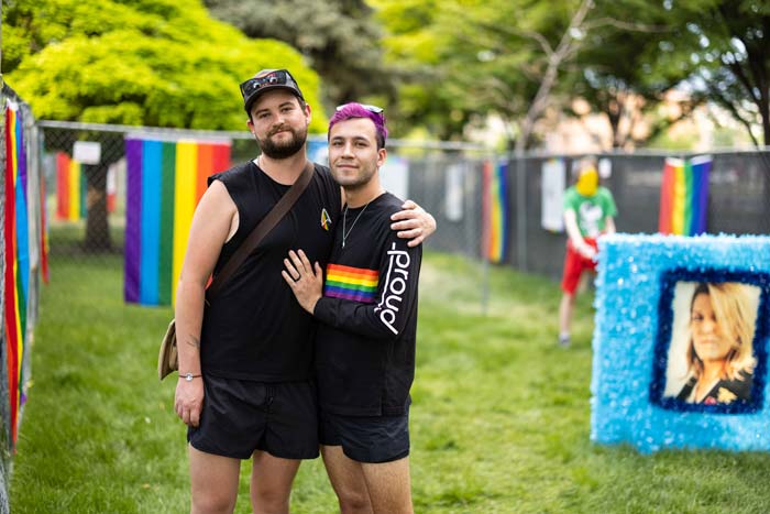 Two men holding each other standing on the exhibit grounds with rainbow flags behind them.