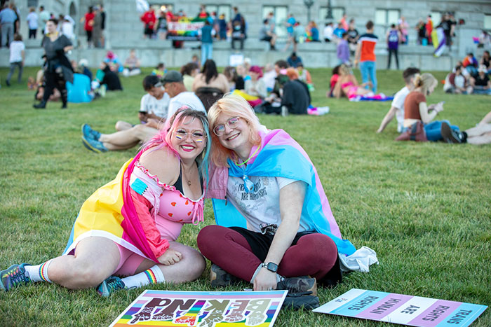 Two people sitting on the grass with others in the background. One has a pansexual flag wrapped around them and the other has a trans flag wrapped around them