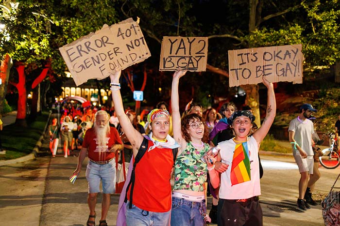 Three people hold signs in front of a crowd. The first says 'Error 404: Gender Not Found...', the second says 'Yay 2 B Gay', and the third says 'It's Okay to be Gay!