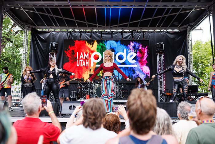 Three drag queens on stage with the 2022 Pride logo in the background and people filming on cell phones in the foreground