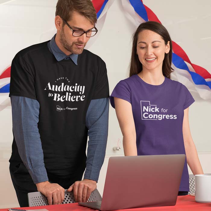 A man on the left wears a black shirt that says 'I have the Audacity to Believe'. A woman on the right wears a purple shirt that says 'Nick for Congress.'