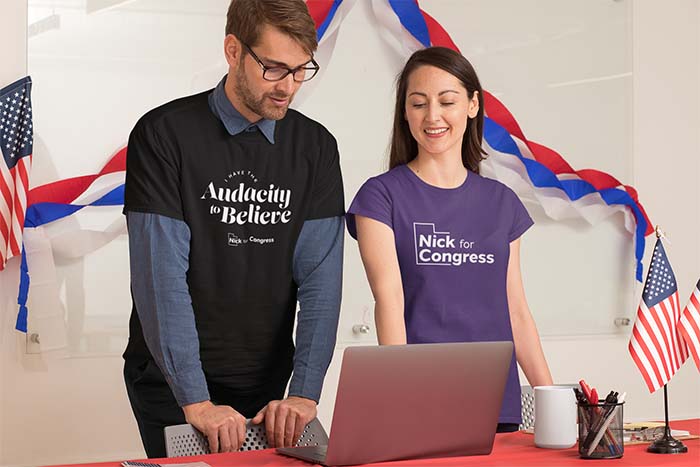 A man on the left wears a black shirt that says 'I have the Audacity to Believe'. A woman on the right wears a purple shirt that says 'Nick for Congress.'