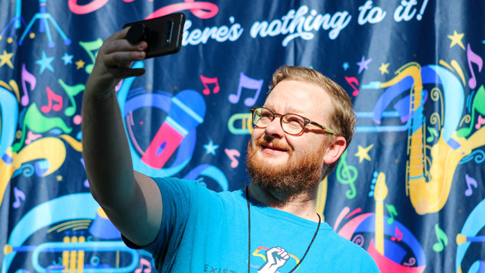 Man taking a selfie in front of a photo backdrop with a blue background and multicolor musical instruments.