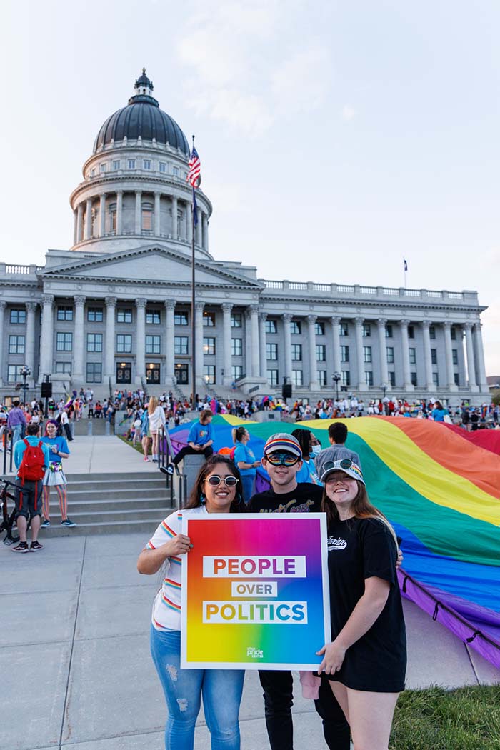 Three people stand in front of a crowd and the Utah Capitol building holding a rainbow sign that says 'People Over Politics'
